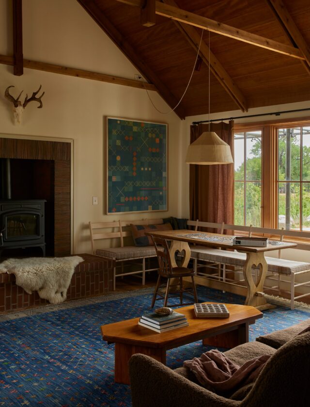 The living room has the only vaulted ceiling in the main house, which creates a moment of punctuation in a floor plan that flows in a meandering and loopy way. Consistent to the rest of the home, we kept the detailing simple, though sometimes those are trickier to coordinate than they imply. These king post trusses, sandwiched between double top chords / rafters, were designed to strike a balance of substantial, rustic weight that doesn’t overtake the room. Getting them right took several studies, conversations, and detail drawings in coordination with the construction team.

One of our favorite elements here is the fireplace. A raised hearth is a great place to perch when folks are gathered, and we surrounded the stove with clay creasing tiles from @northernrooftiles (they clad the fireboxes in this room and the kitchen, too). We also designed a log holder with the talented @slmetalworks that sits nearby.

We commissioned a glowing collage from @lenawolffstudio via our pal @sarahshepardgallery, which hangs above a large corner bench from @brianpersico. This art is in conversation with a @geesbendquiltmakers quilt on the opposite wall. Underneath it all, we laid a lapis blue rug from @_salamhello_  that we think ties everything together in a lighthearted, cheerful manner.

Just outside are a dining table and sectional, accessed by the kitchen or guest bedroom. This nook is the one area of the home that is (mostly) protected from intense winds that can blow through without warning. We framed the area with a custom steel pergola that will eventually host plants which provide additional coverage for the calm and shaded living room.

We have a lot of process documentation to share from our friends who contributed to this room, so keep your eyes on our stories if you’re curious!

The link to the feature is in our bio! 🏔️

Interior Architecture & Design: @landedinteriors #landedinteriors
Project Team: @heathermenegat, @rebeccamullerdesigns, Tiffany Kim, Luna Sibai, Laura Marx, Hanna Dooley
Architecture & Construction: @coburnpartners 
Landscape: @treeoflifelandscaping 
Photography: @yoshihiromakino 
Styling: @rainakattelson 
Publication: @archdigest